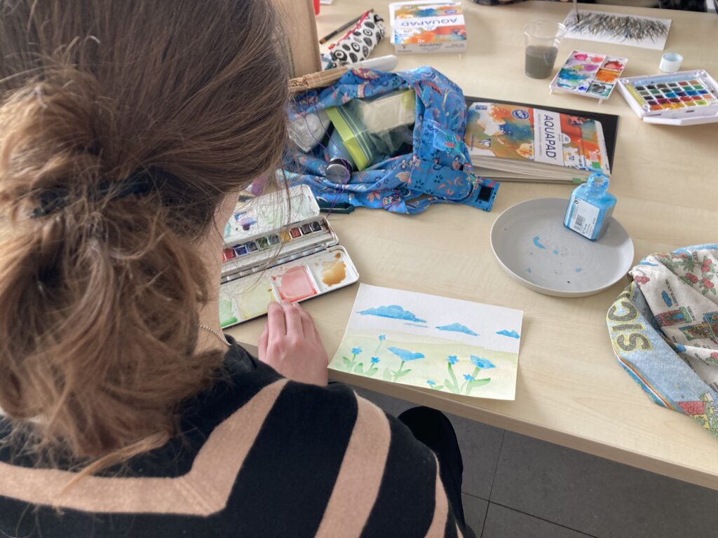 A young woman taking an art class, seen from behind, as she paints a delicate mountain landscape in watercolor in a small sketchbook.