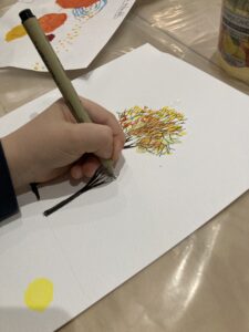 A close-up of a child's hand drawing a tree with a fine-tip marker (fineliner) on top of an autumn-themed watercolor background.