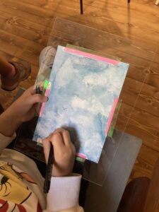 A child painting a cloudy sky using the wet-on-wet watercolor technique on a sheet of paper secured with colored tape.